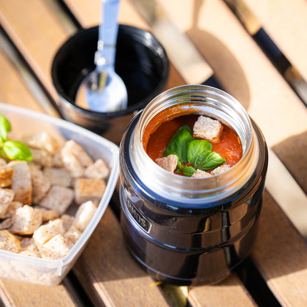 An opened thermos flask reveals fresh tomato soup with basil and croutons. Next to the thermos is a container with extra croutons and a spoon.