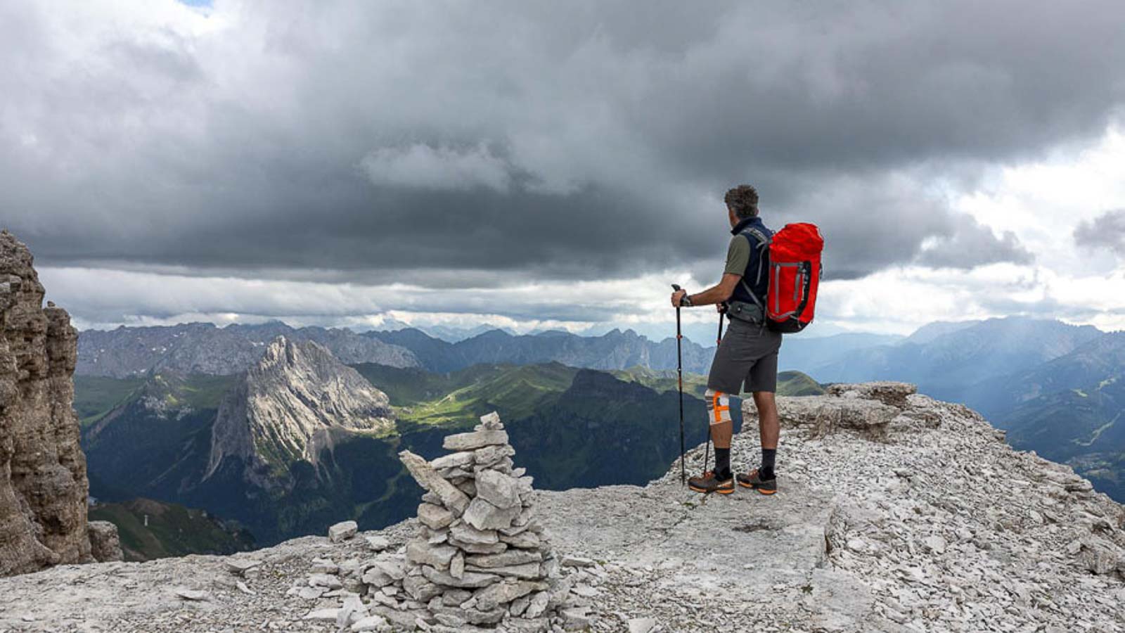 A person with a red backpack stands on a rocky ledge in the Dolomites, gazing into the distance. The sky is cloudy, and there is a cairn structure in the foreground.