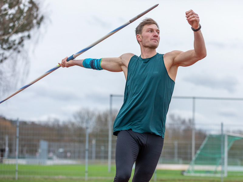 Thomas Röhler holds a javelin in his hand and is about to throw it. He wears an elbow brace and is on the training ground.