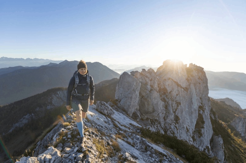 A hiker wearing Outdoor Merino Compression Socks walks on a narrow mountain trail at sunrise. These compression socks provide optimal support and comfort for challenging hikes.