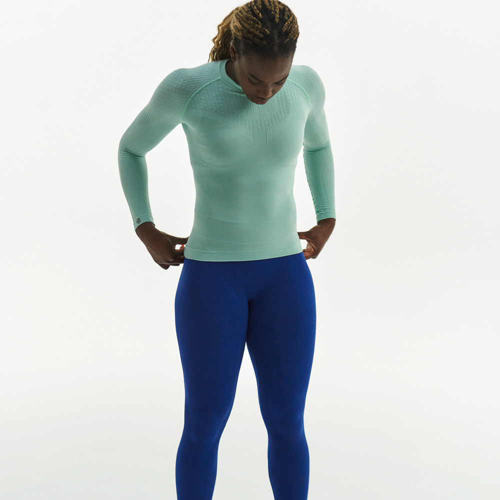 Woman in mint green compression top and dark blue compression trousers looks at the floor and has her hands on her hips