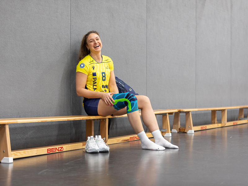 A player of SSC Palmberg sits on a bench in a sports hall, laughing at the camera. She wears a knee brace.