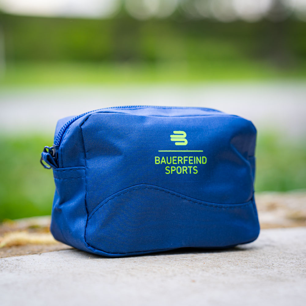 A small, blue cosmetic bag from Bauerfeind Sports with a zipper sits on a concrete surface outdoors. The bag displays the Bauerfeind logo and is compact for on-the-go use.