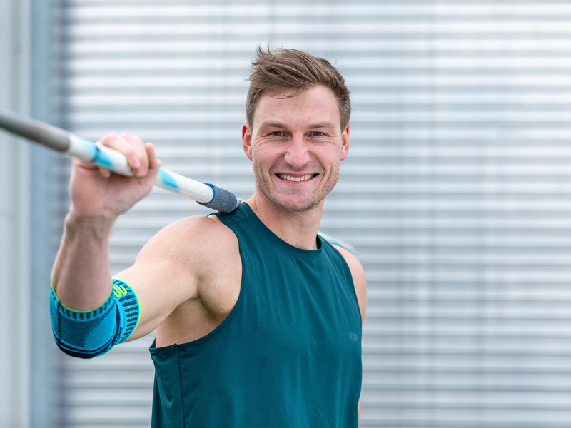 Thomas Röhler stands in front of a goal net holding a javelin on his back, wearing an elbow brace.