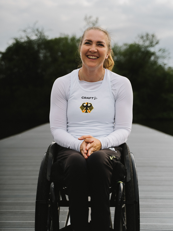 The German athlete Edina Müller sits in a wheelchair on a dock and laughs at the camera.