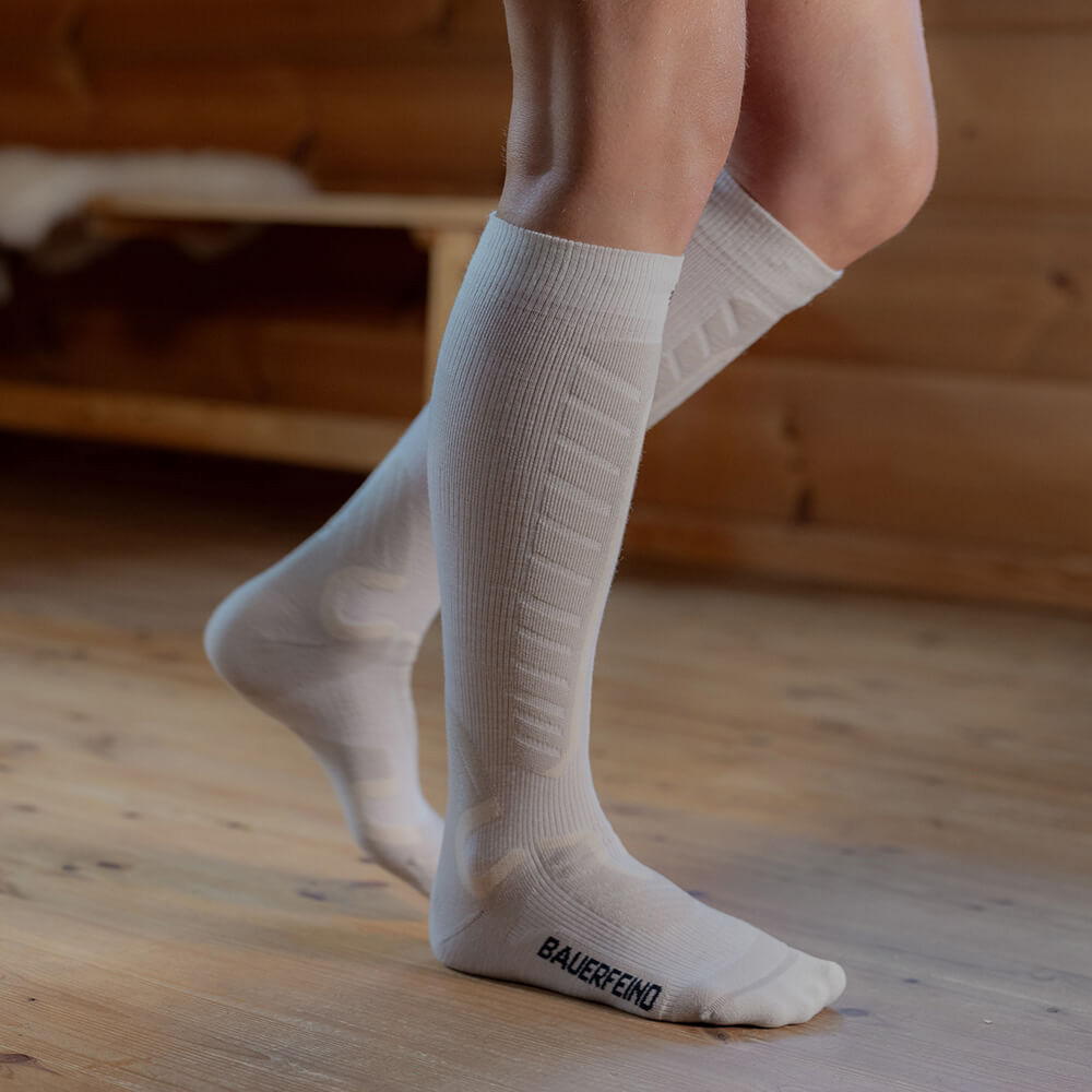 Close-up of white compression ski socks that reach just below the knees. The person stands on a wooden floor in a cabin. The socks offer extra comfort and support with their padded zones for skiing.