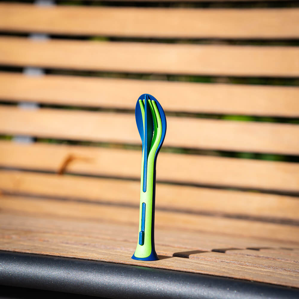 A green and blue cutlery set stands upright on a wooden bench. The set consists of a knife, fork, and spoon, which are compactly stacked together.