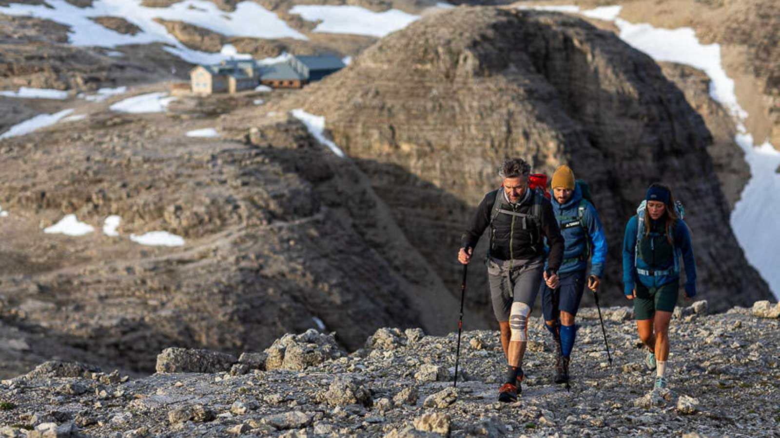 Rocky landscape in the Dolomites, with a group of hikers in the foreground wearing hiking bandages and socks, and a mountain hut in the background.