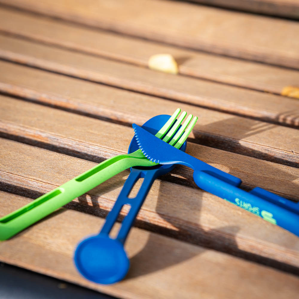 A green and blue cutlery set lies crossed on a wooden bench. The set is ideal for outdoor meals and compact packing.