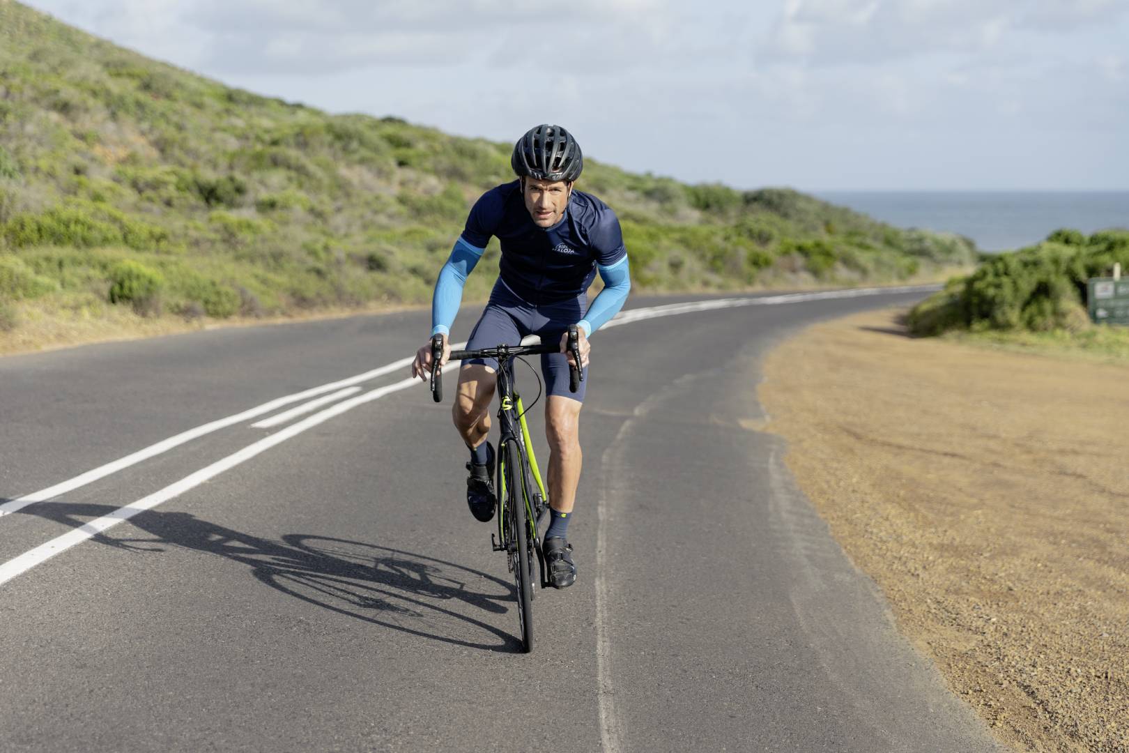 A man is riding a racing bike on a coastal road in cloudy weather. He is pedalling in the cradle.