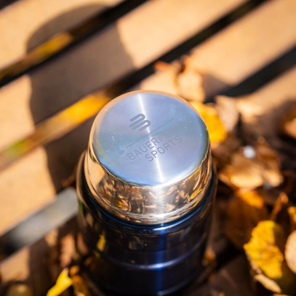 Close-up of the shiny lid of a thermos flask engraved with the Bauerfeind Sports logo. The lid rests on a wooden bench, surrounded by autumn leaves.