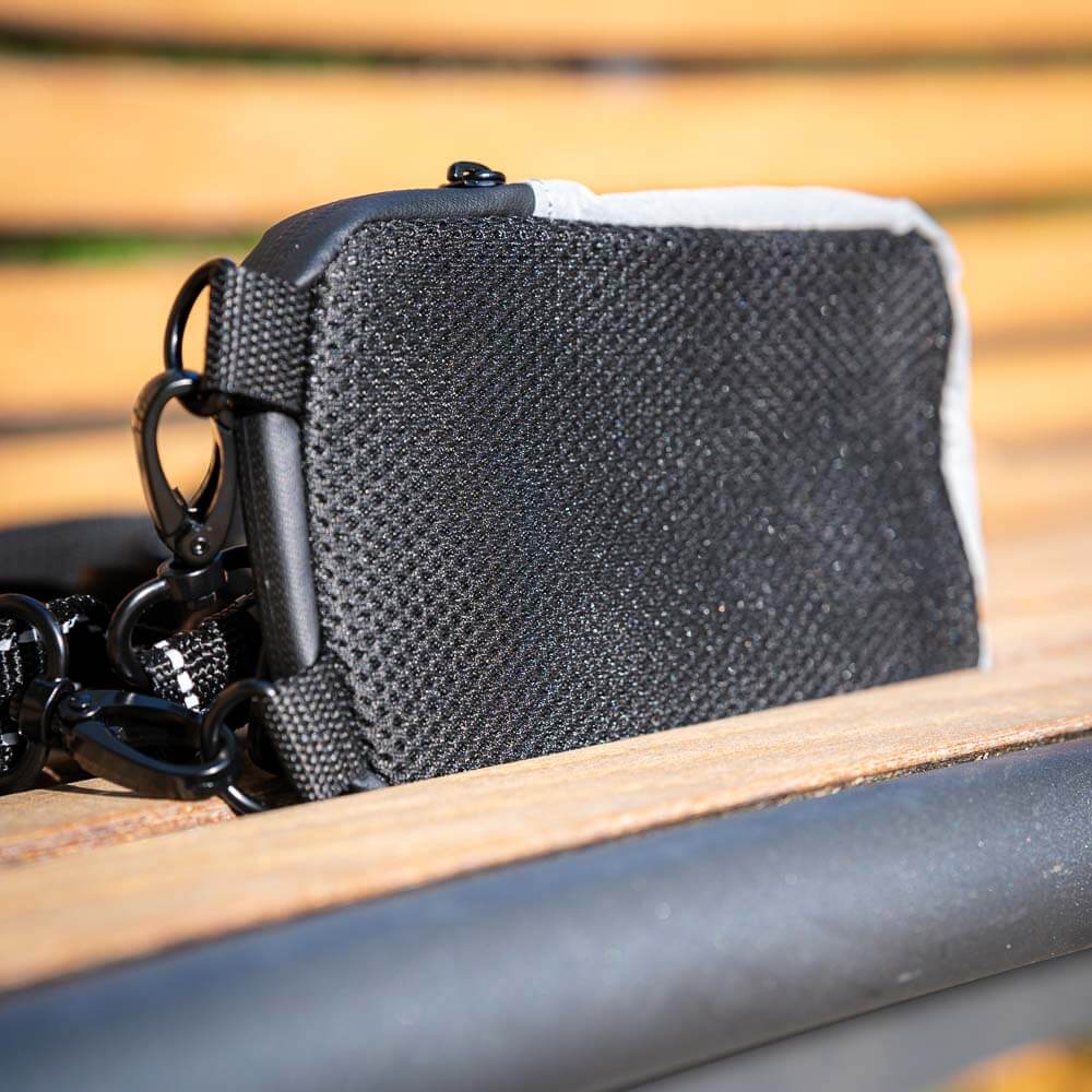 Close-up of the back of the gray Bauerfeind Sports pouch, featuring a breathable black mesh material. The pouch rests on a wooden bench and has two carabiners for attachment.