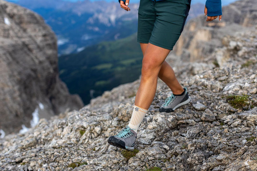 A hiker navigates rocky terrain with outdoor shoes and an orange ankle brace. A vast mountain landscape stretches out in the background.