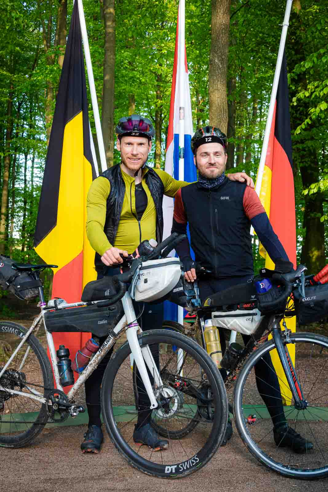 Two cyclists stand at the border triangle of Germany, Belgium and the Netherlands and laugh into the camera.