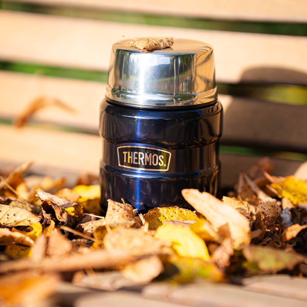 A blue thermos flask sits on a wooden bench surrounded by autumn leaves. A small leaf rests on the shiny lid. The flask features the "Thermos" logo and shines in the autumn setting.