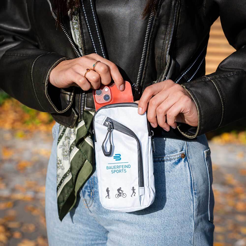 A person stores a smartphone in the gray Bauerfeind Sports pouch worn on the hip. The pouch is compact and ideal for outdoor activities.