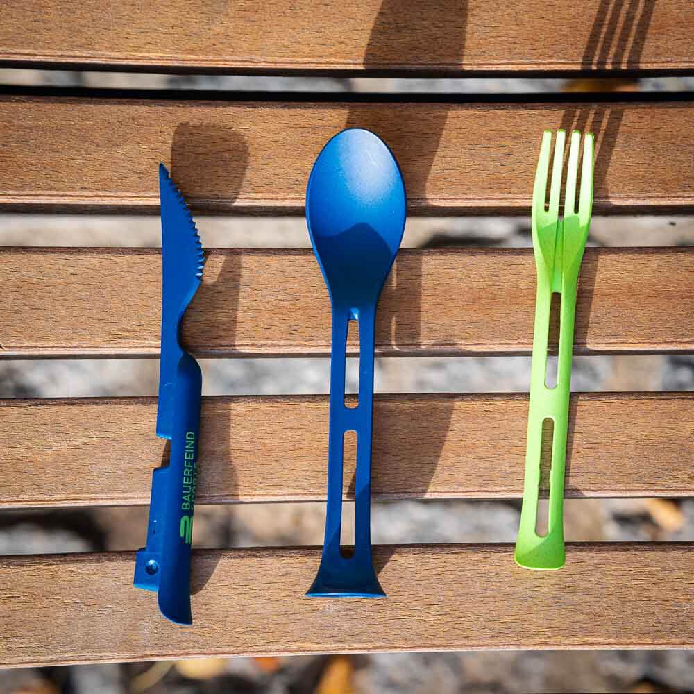 A disassembled cutlery set with a blue knife, blue spoon, and green fork laid side by side on a wooden bench.