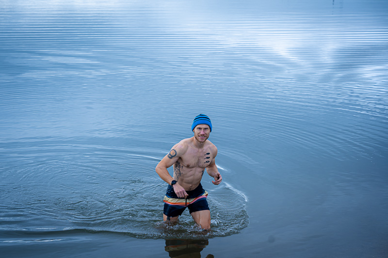 A man wades out of the lake, filmed while ice bathing in swim trunks and a blue beanie in the autumn setting.