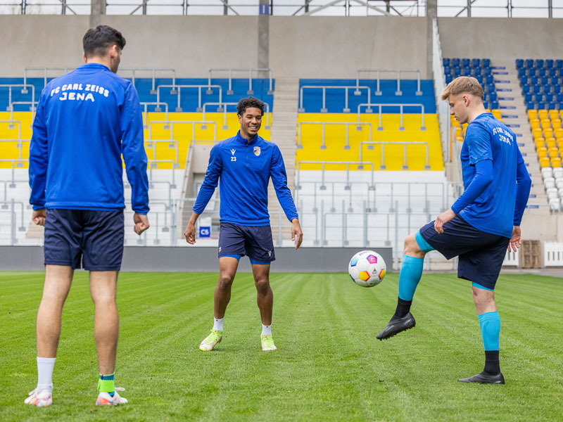 Three players of FC Carl Zeiss Jena in the stadium training with a football, wearing various Bauerfeind Sports products