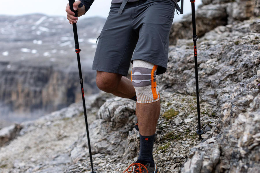 A hiker with trekking poles and an orange knee brace walks on a rocky mountain path. In the background, rugged cliffs and misty mountain landscapes are visible.