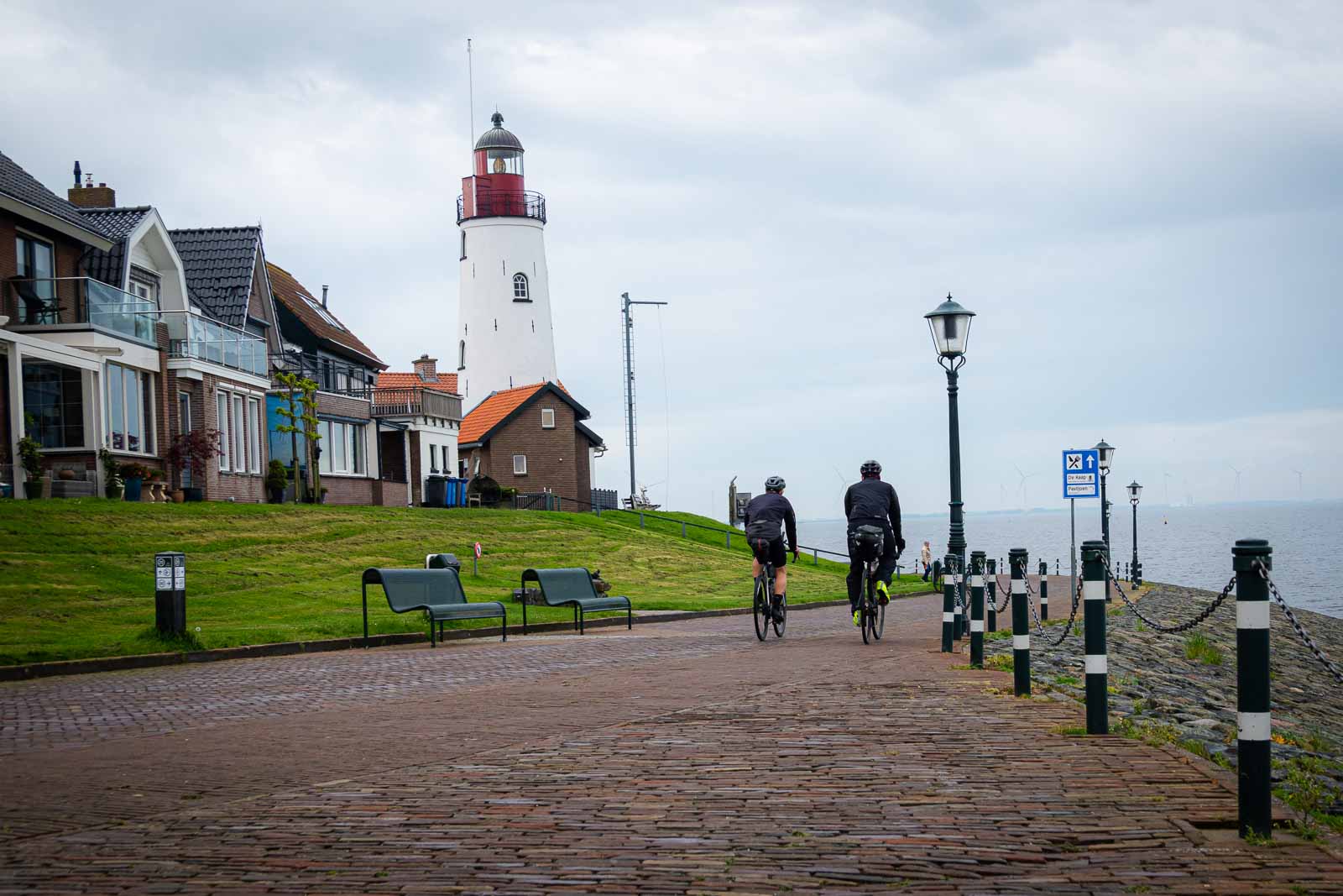 Two ultra-cycling cyclists from behind riding along a promenade where a lighthouse stands.