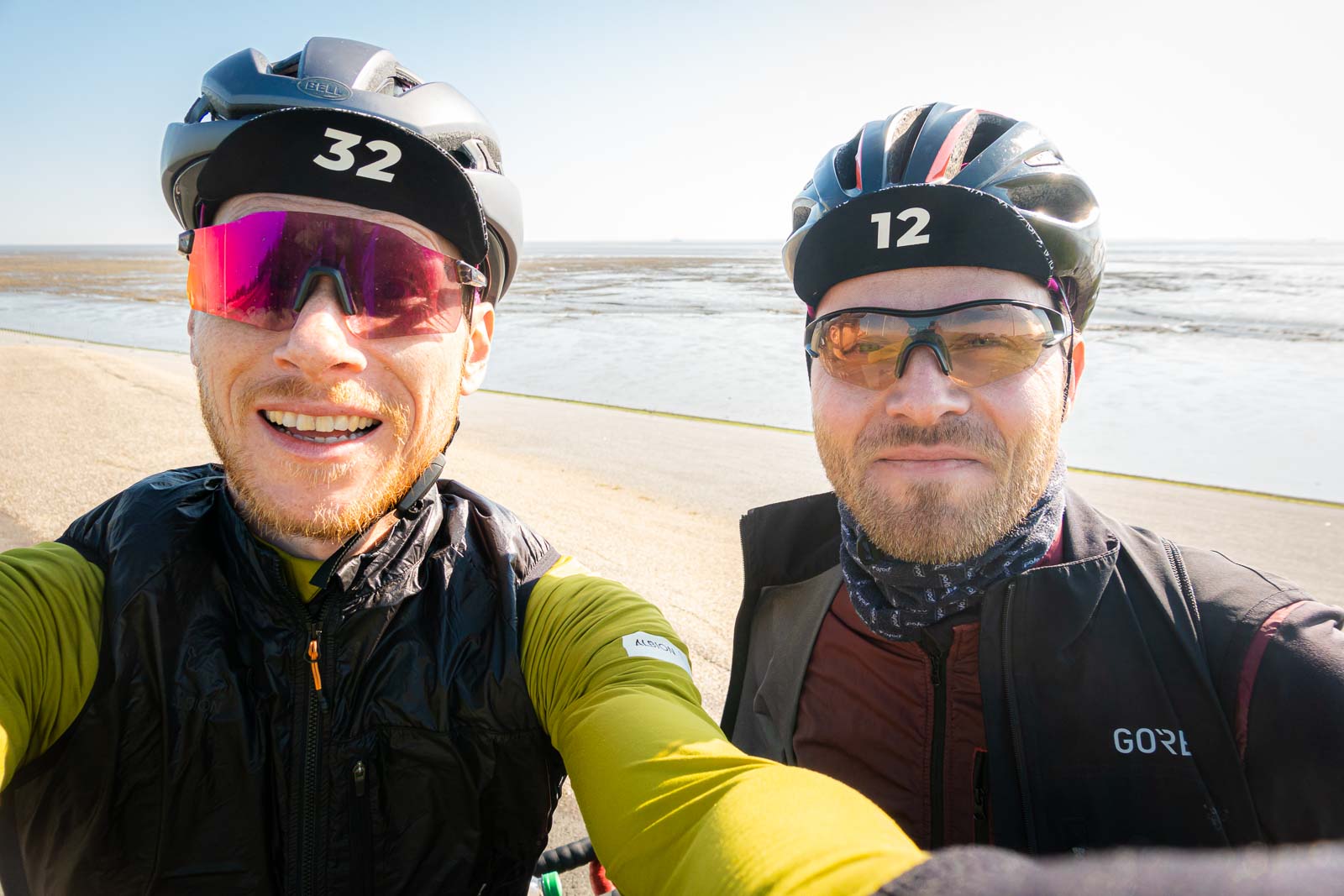 Two cyclists take a selfie of themselves while standing in front of a beach panorama and laughing.